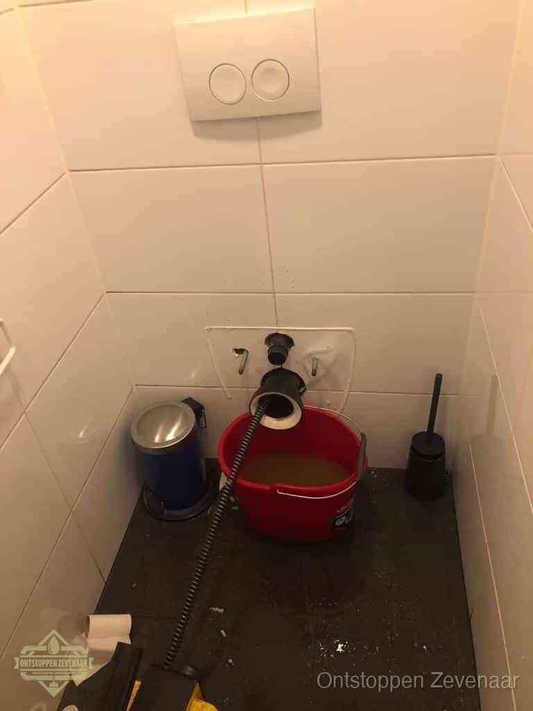 Een ontstopte wand-wc met verwijderde toiletpot waar een ontstoppingsveer in de wanduitgang verdwijnt, het achterhalen waar de verstopping zit in Zevenaar