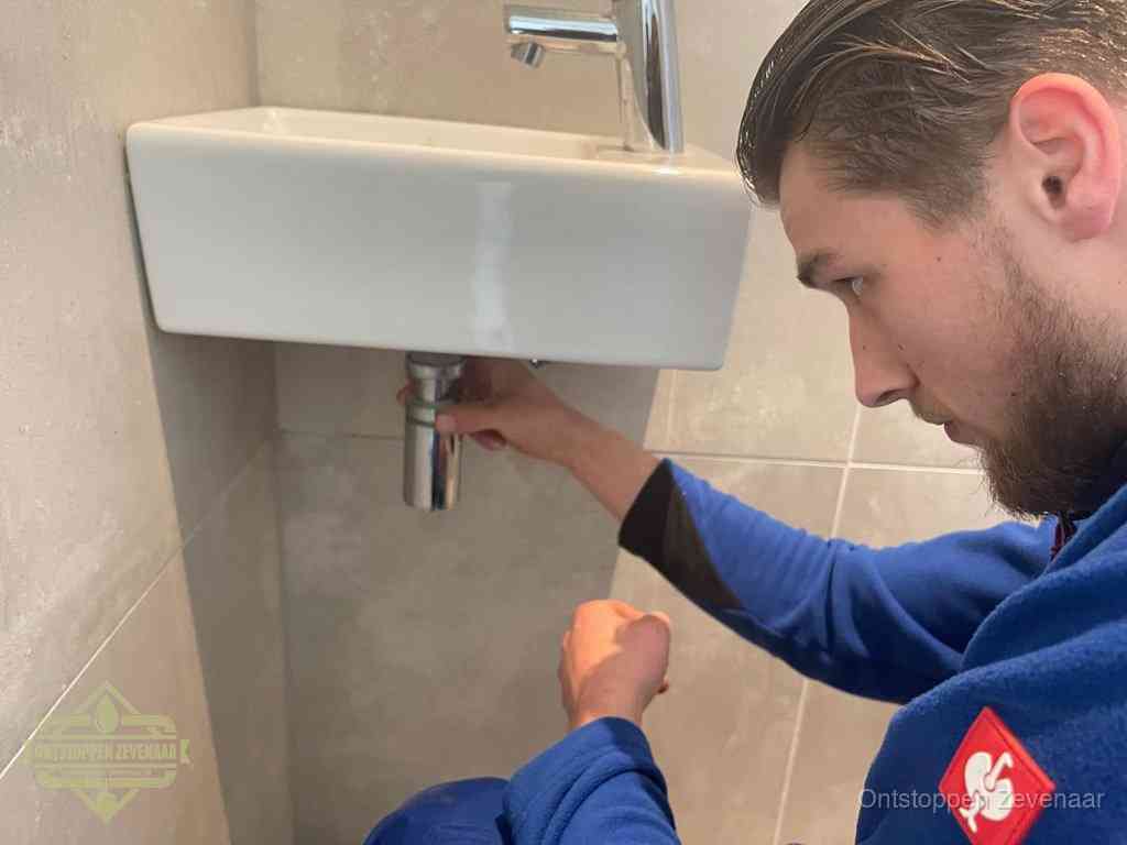 Een monteur draait met de hand de zwanenhalssifon onder een fonteinwasbak in de toiletruimte los om de bron van rioollucht in Zevenaar te vinden