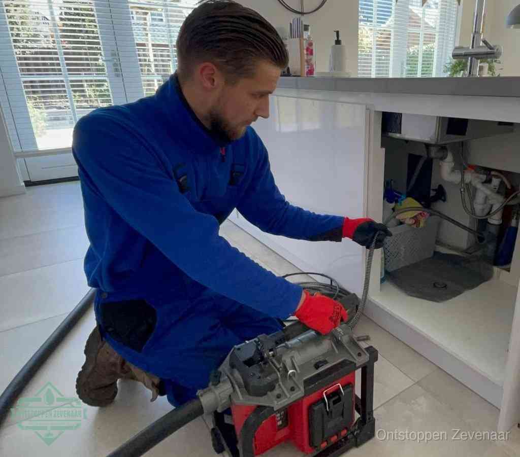 Een monteur met rode handschoenen knielt in een keuken en voert een elektrische ontstoppingsveer in tijdens het professioneel afvoer ontstoppen in Zevenaar
