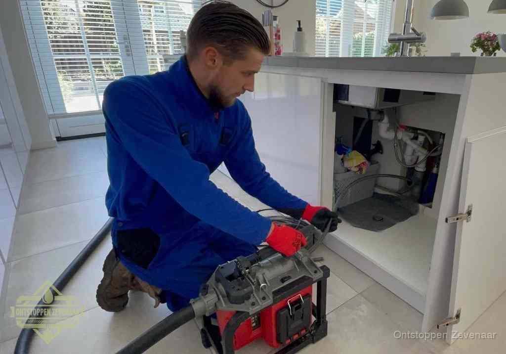 Een monteur voert een professionele elektrische ontstoppingsmachine in de afvoer onder de spoelbak tijdens het gootsteen ontstoppen in Zevenaar